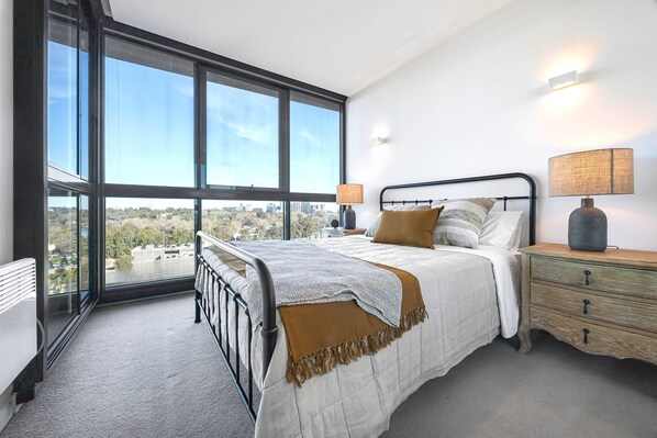 A bedroom with a large window, a bed with a white comforter, a wooden nightstand, and a lamp. - 108 Flinders - Stunning Views & Walk Everywhere - Steps from Fed Square (Melbourne)