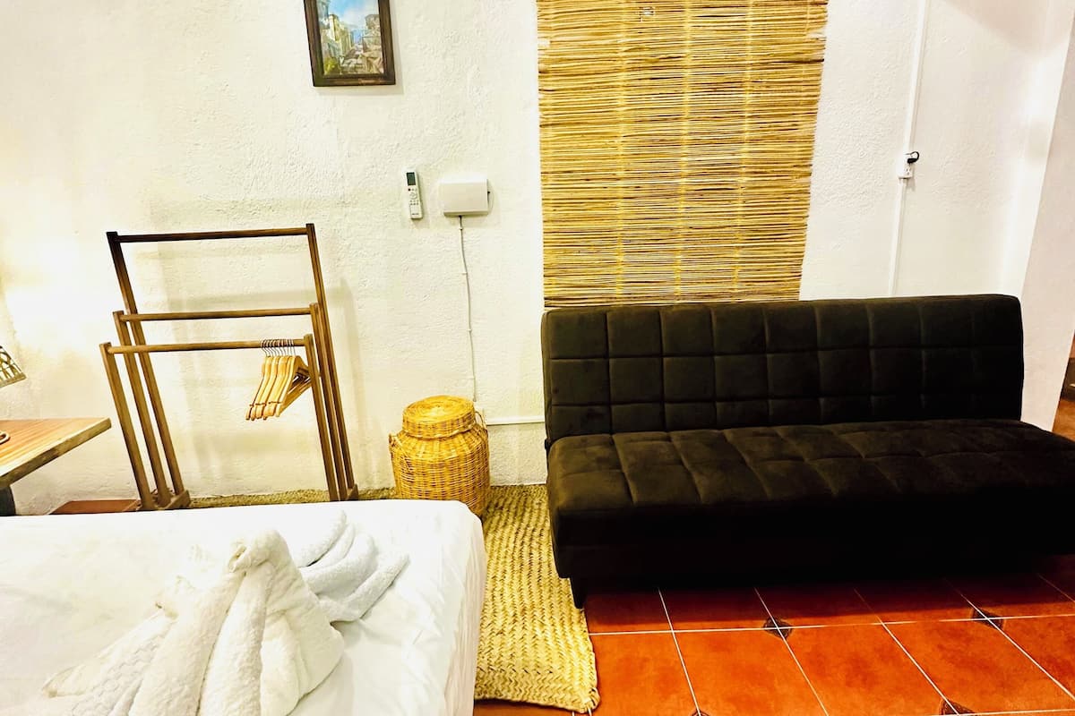 A hotel room with a bed, a black sofa, a wicker chair, a small table, and a television mounted on the wall.