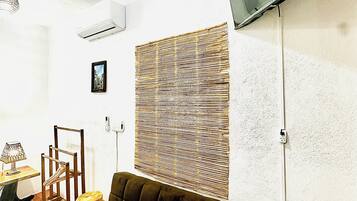 Una sala de estar con un sofá oscuro, un televisor montado en la pared, un sillón de mimbre, una mesita con una lámpara y una alfombra tejida.