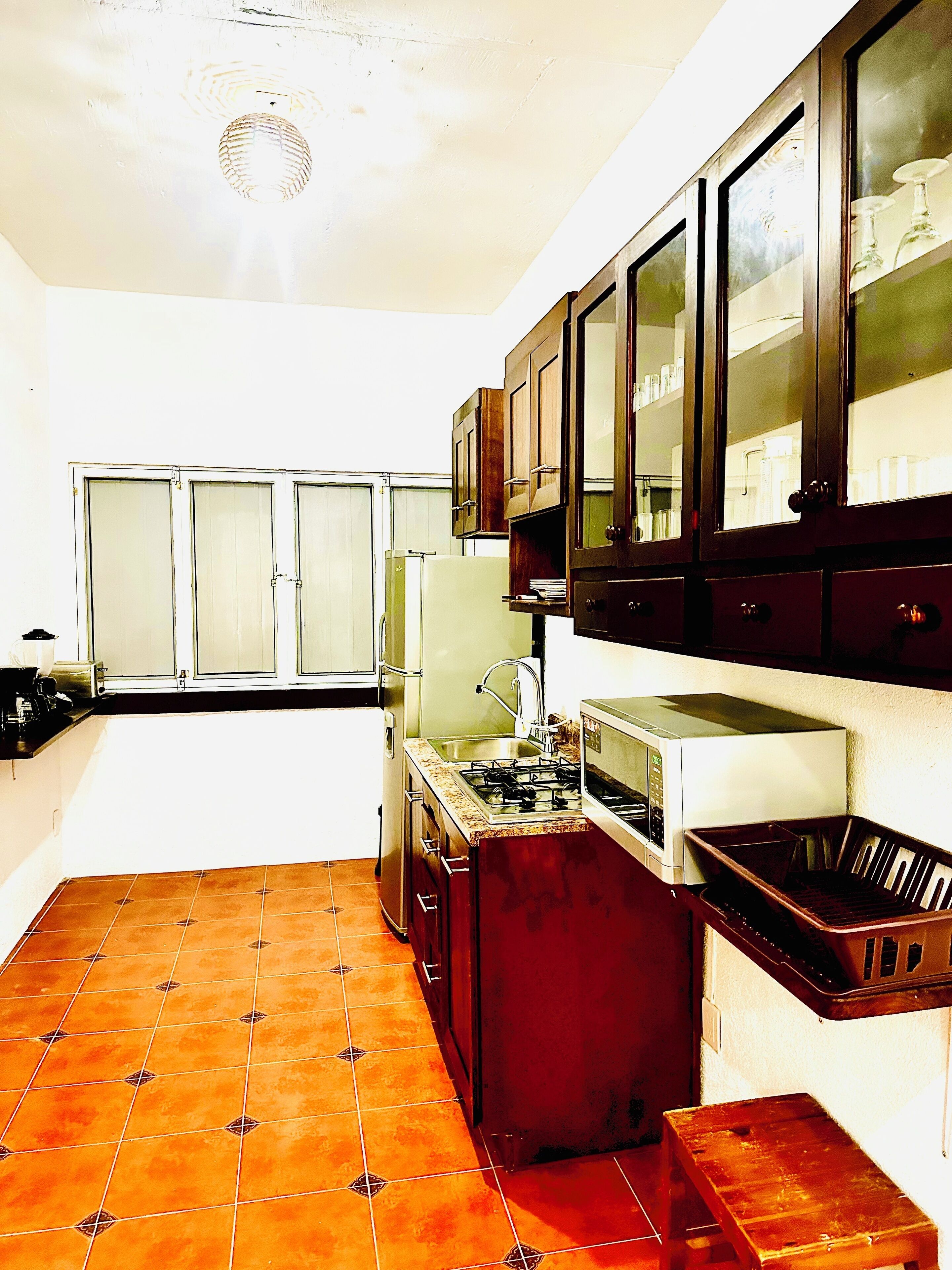 A compact kitchen with a gas stove, a sink, and a microwave. There are cabinets above the counter and a window with blinds.