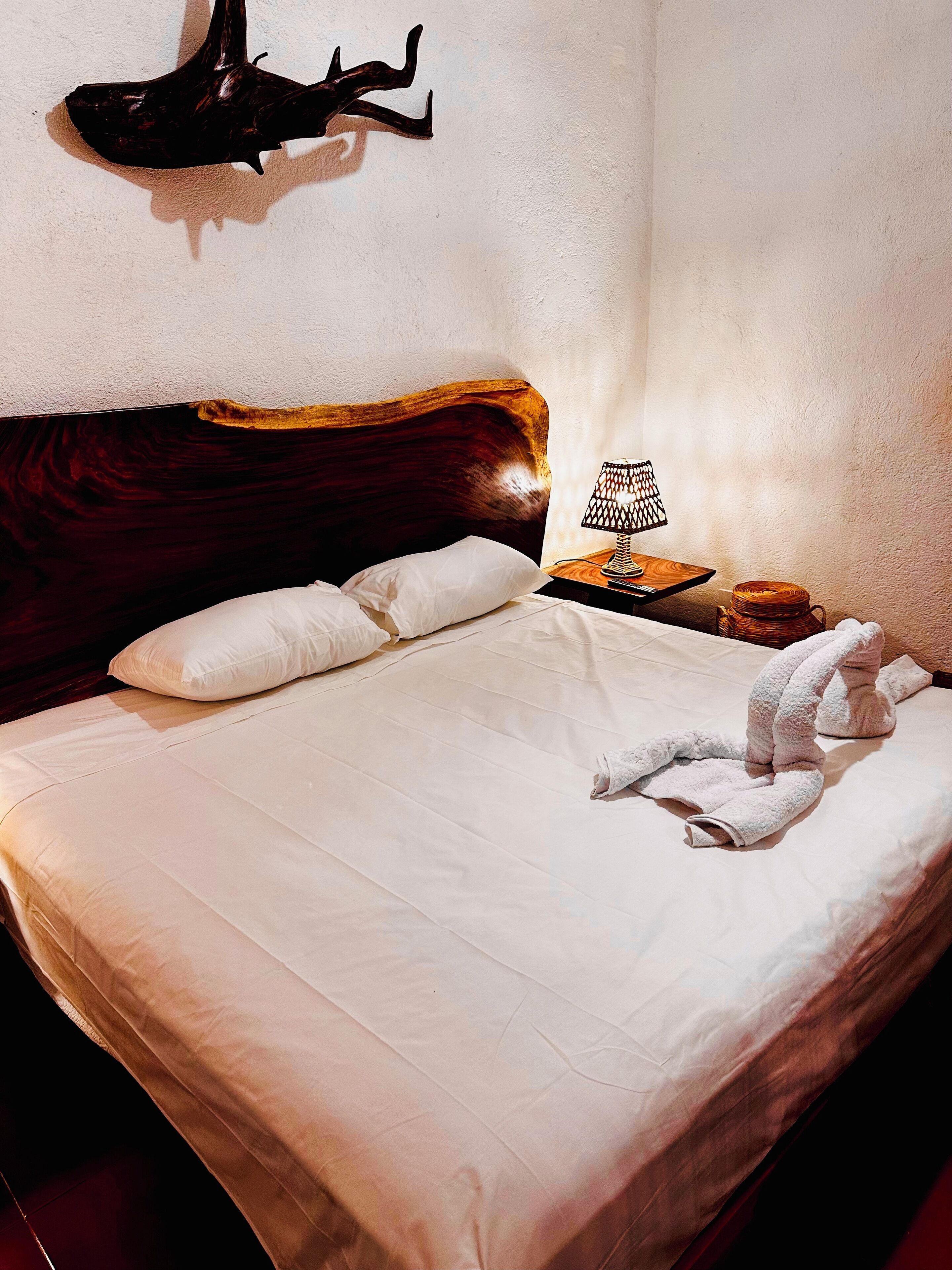Una cama bien hecha con sábanas blancas, cabecera de madera y una escultura de tiburón en la pared.