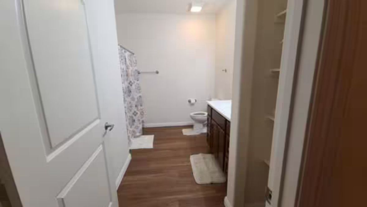 Un pasillo estrecho con piso de madera que conduce a un baño con ducha e inodoro.
