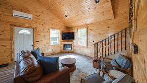 A cozy log cabin interior with a leather sofa, two armchairs, a coffee table, a fireplace, a TV, and a staircase. - Cabin w/ River Boat Ramp~Private HotTub~Sauna~Lake Cumberland~Firepit~Pickleball (Jamestown)