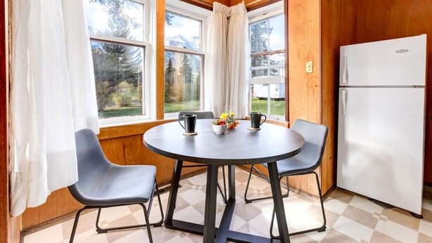 A compact kitchen with a round table, two chairs, a refrigerator, and large windows with curtains. - Huckleberry Lodge Mountain Getaway Near Banff! (Harvie Heights)