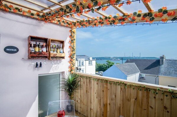 A balcony with a wooden pergola, a wine bottle shelf, and a view of the sea and buildings.