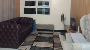 A living room with a dark brown sofa, a light-colored armchair, a beige bench, and a patterned rug. There is a ceiling light fixture and a window with blinds. - Chic 1-bedroom building in fantastic Pasay City with AC, fitness room (Pasay City)