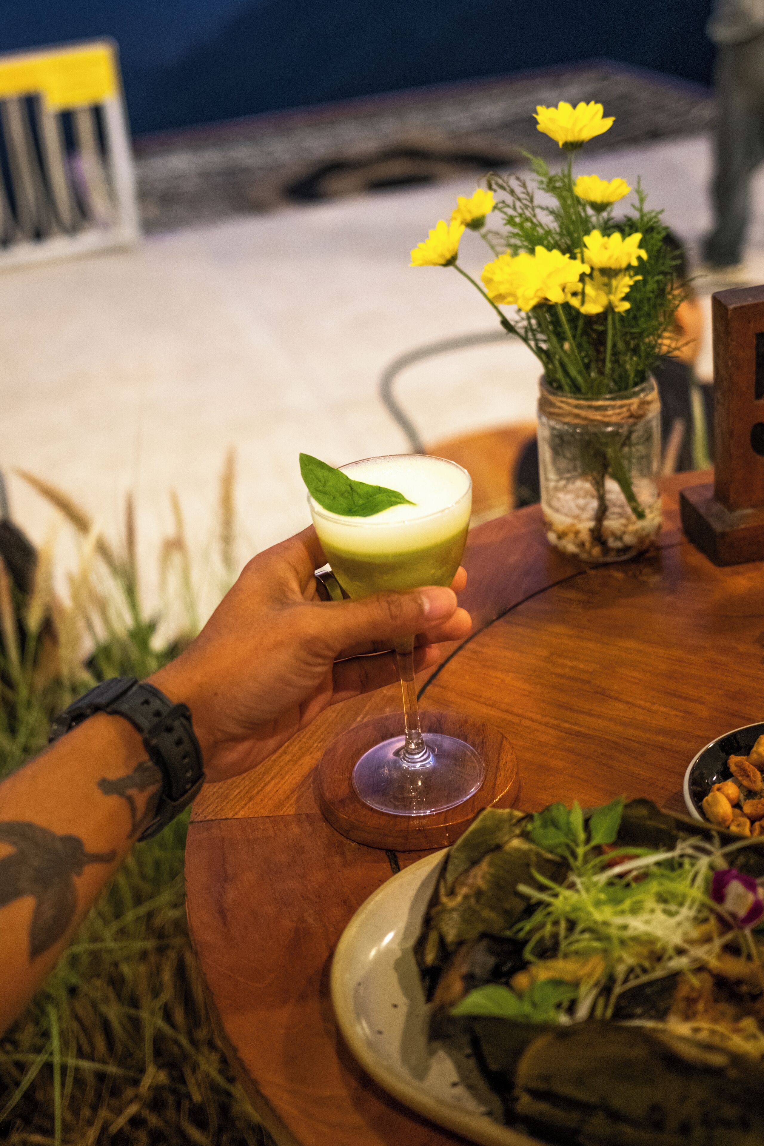 A hand holding a cocktail glass with a green drink, a plate with food, and a vase with yellow flowers on a wooden table.