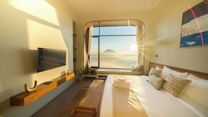 Habitación de hotel con un ventanal que ofrece vistas a montañas y nubes, un televisor de pantalla plana en la pared, una mesita de noche de madera y una cama con ropa blanca y almohadas estampadas.