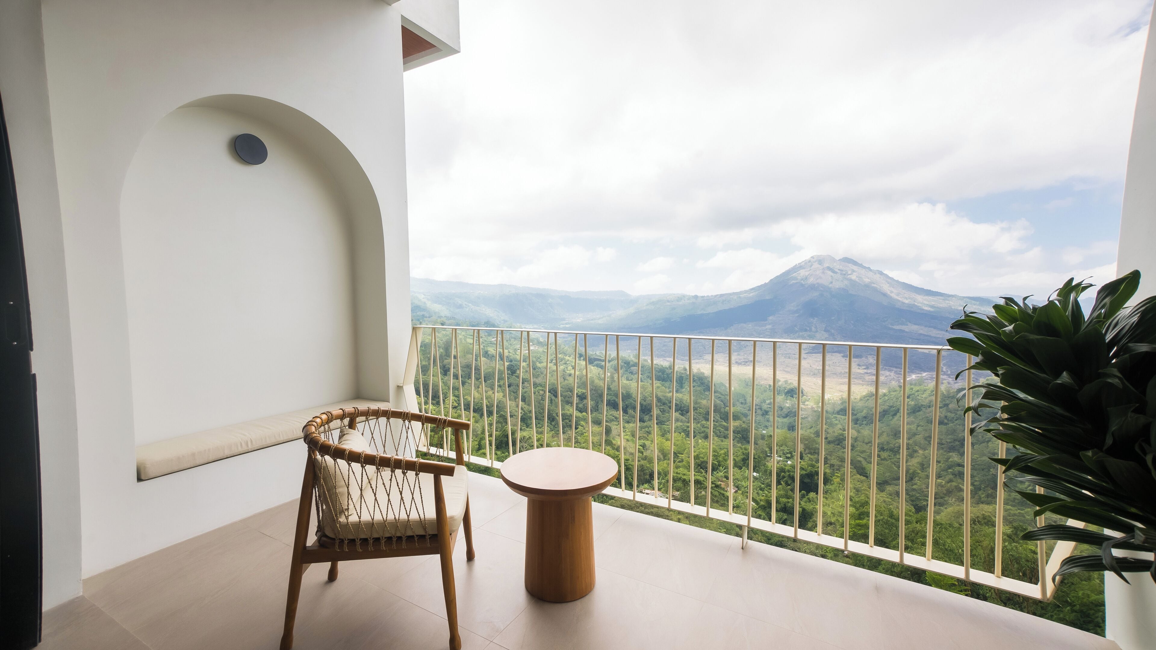 A balcony with a wicker chair and a small round table, overlooking a mountainous landscape.