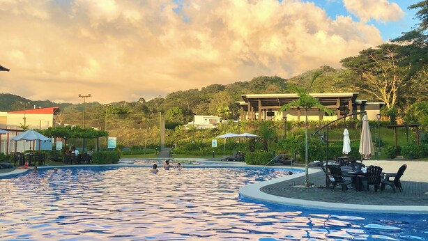 A swimming pool with lounge chairs and umbrellas, surrounded by greenery and buildings. - Family condo with resort-style pools, close to beaches, nature and adventure. (puntarenas)
