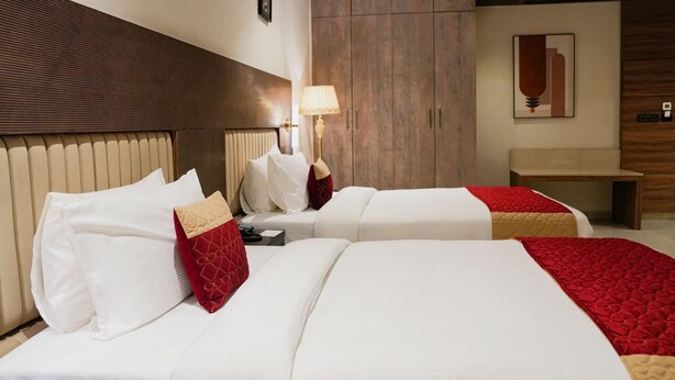 A hotel room with two beds, a wooden headboard, a bedside table with a lamp, and a wooden wardrobe. - The Meriton (New Delhi)