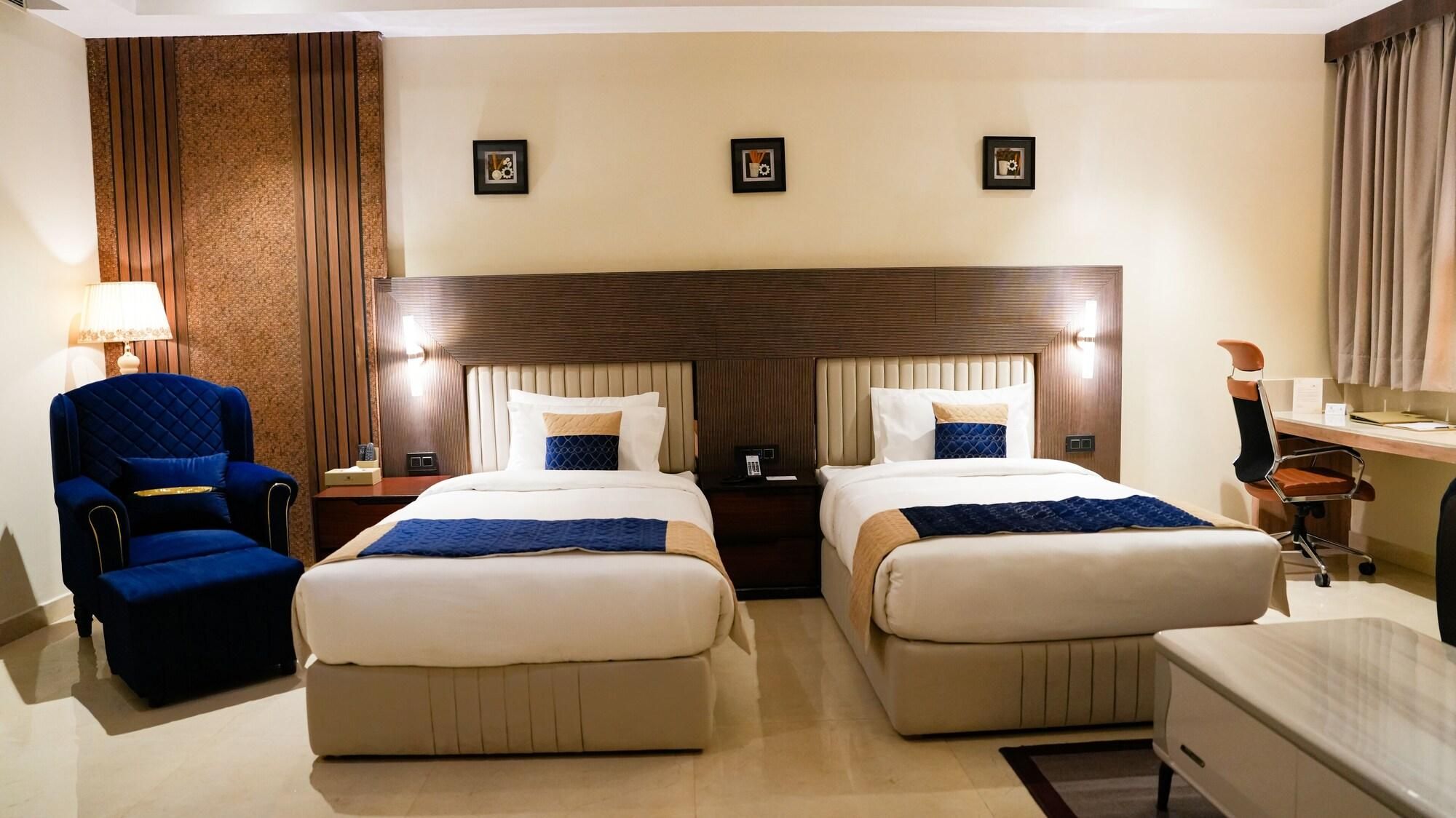 A hotel room with two beds, a chair, a desk, and a lamp.