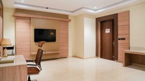 A modern hotel room with a flat-screen TV mounted on a wooden wall, a wooden door, and a desk with a lamp. - The Meriton (New Delhi)