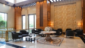 A spacious lobby with a central round table, multiple seating areas, and a large window providing natural light. - The Meriton (New Delhi)