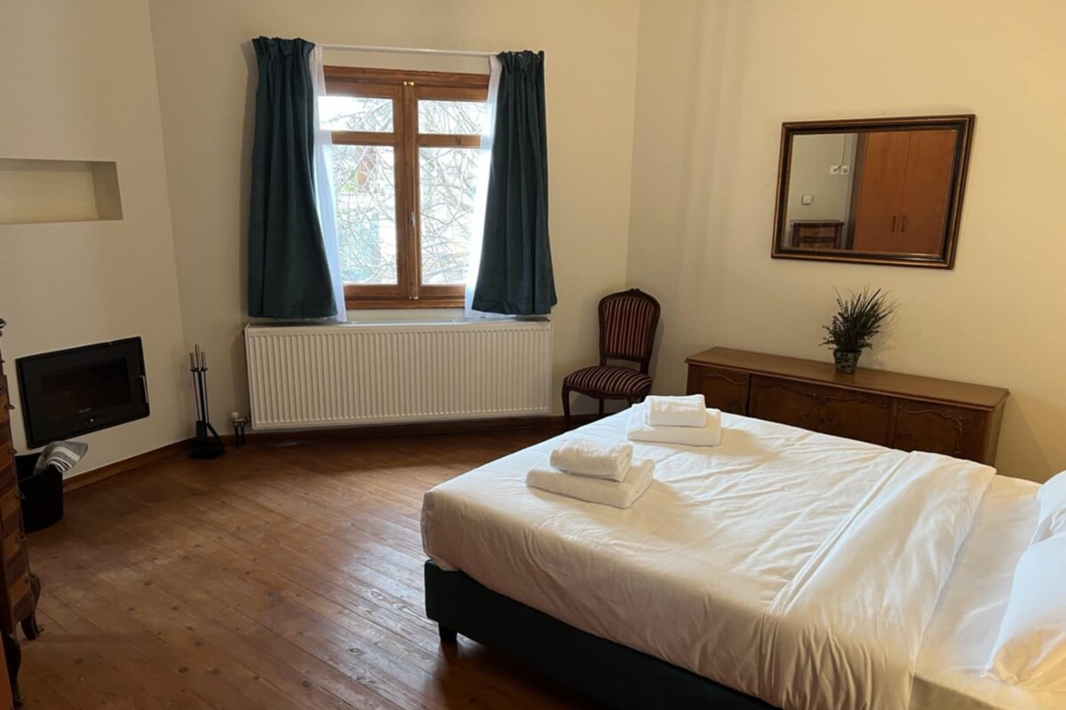 Una camera da letto con un letto, una cassettiera, una sedia, un termosifone, una finestra con tende e uno specchio a parete.