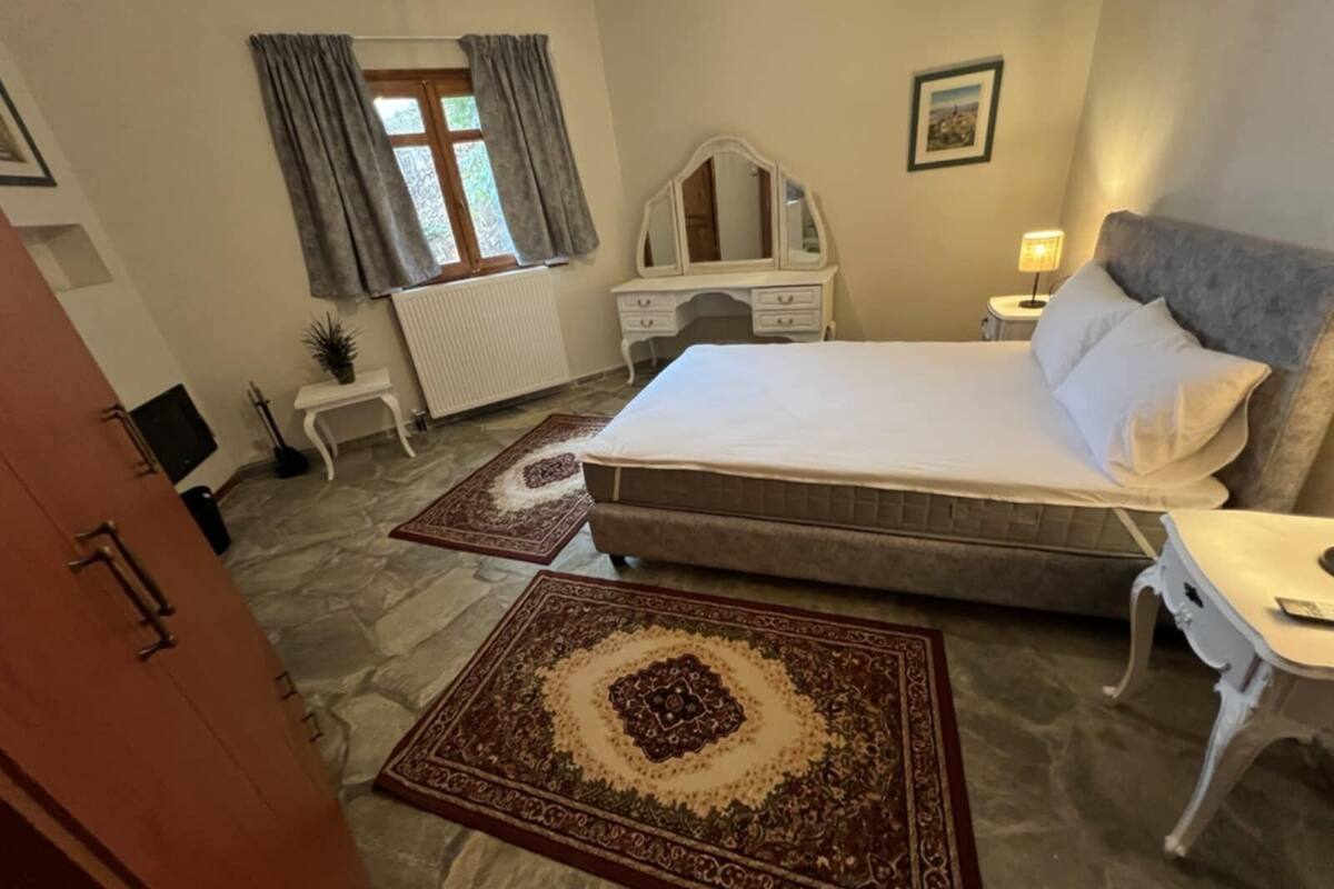 A bedroom with a large bed, a dresser, a nightstand, a lamp, and a window with curtains.