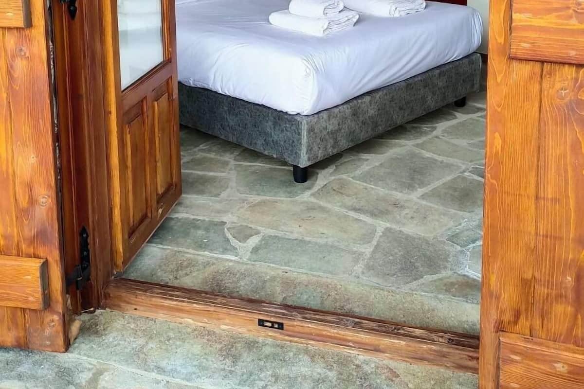 A bedroom with a stone floor, a bed with white linens, and a nightstand with a lamp.