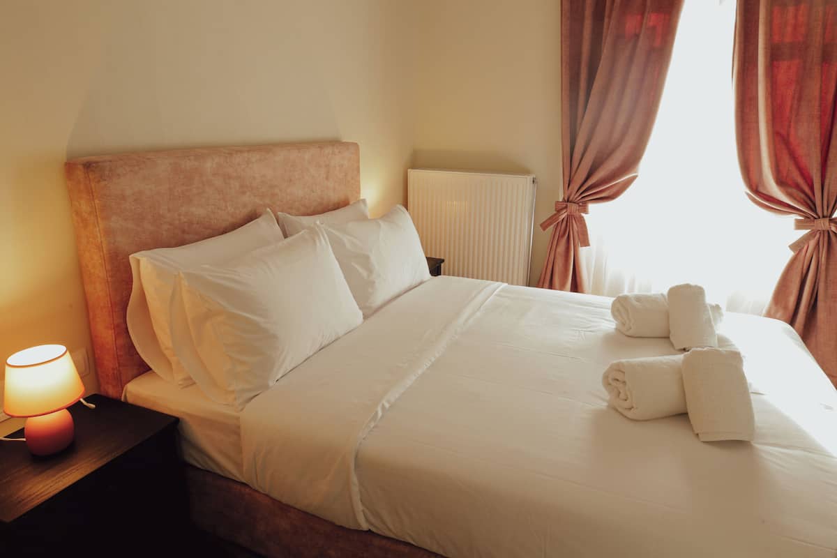 A neatly made bed with white linens and pillows, a bedside lamp, and a window with curtains.