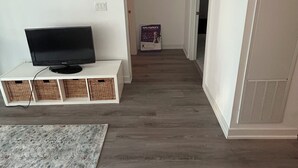 Un couloir avec un téléviseur posé sur un meuble blanc, des paniers en osier, une affiche et une grille d’aération sur le mur.