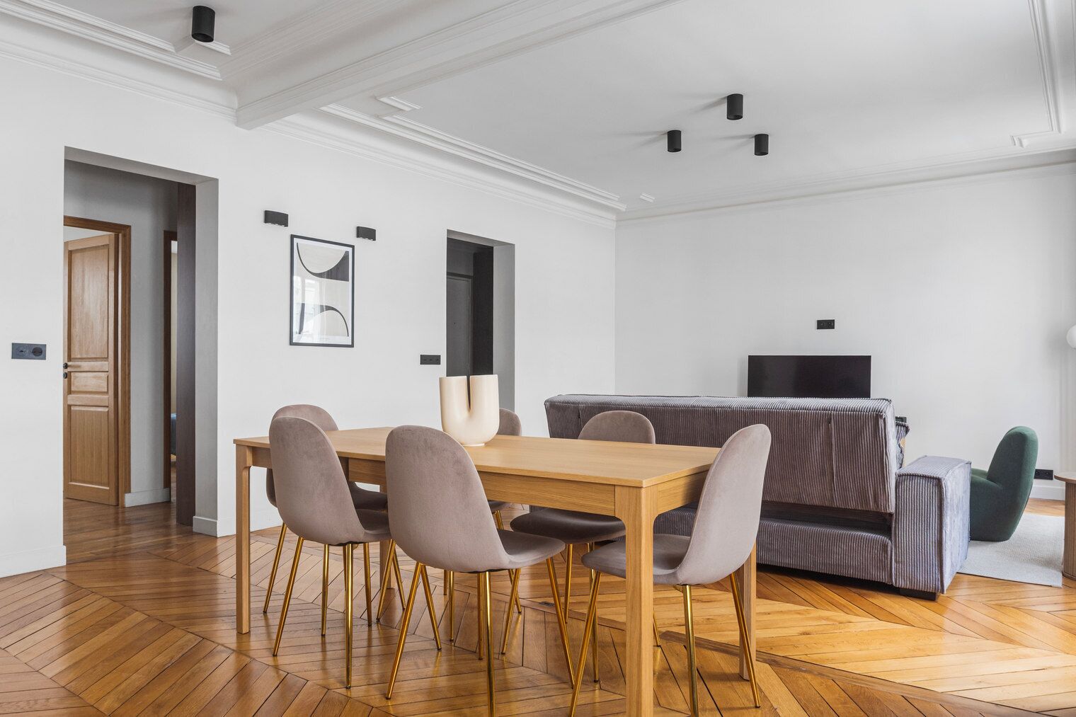 A modern living room with a wooden dining table, four chairs, a grey sofa, and a television.