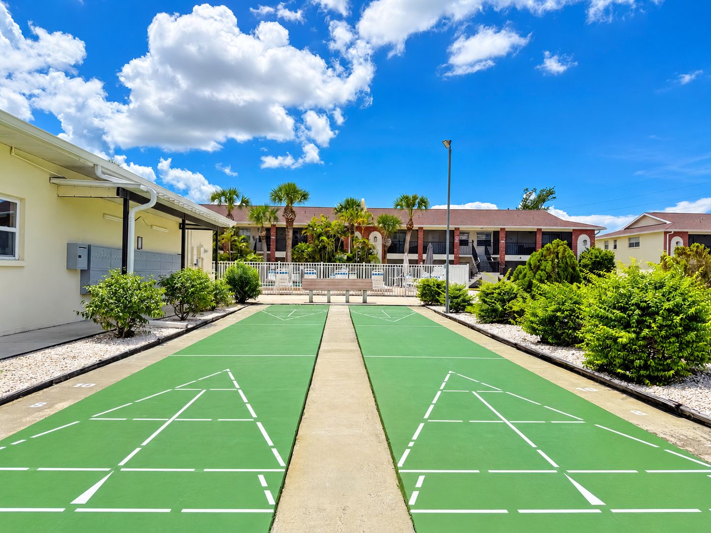 Ein Innenhof mit zwei grünen Shuffleboard-Plätzen, einem weißen Zaun und einem Gebäude mit zahlreichen Fenstern und Palmen.
