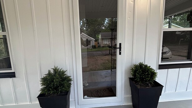 Une porte d’entrée avec un paillasson sur lequel est écrit « bonjour », deux plantes en pot et une fenêtre sur le côté droit.