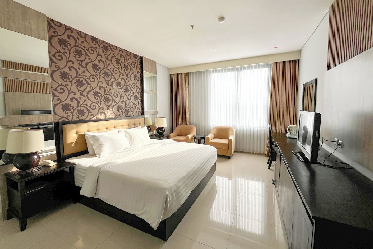 A hotel room with a large bed, two armchairs, a desk with a computer, and a TV.
