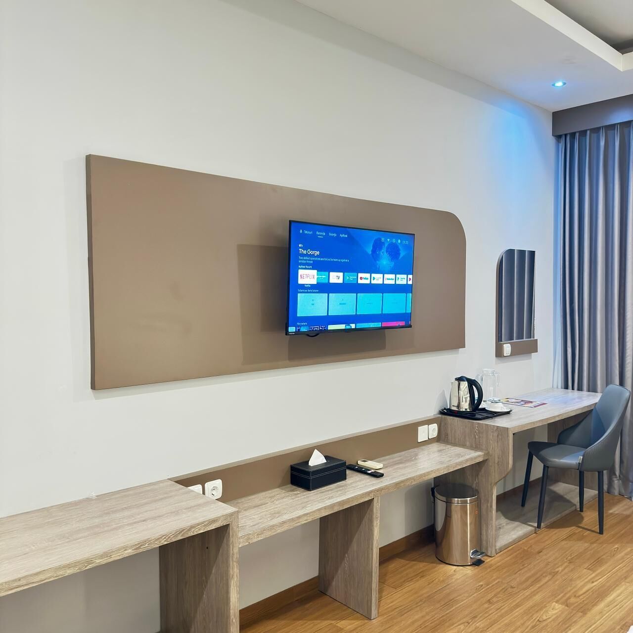 Habitación de hotel moderna con televisión de pantalla plana montada en la pared, un escritorio de madera con un bote de basura, una silla y una pequeña mesa con una cafetera.