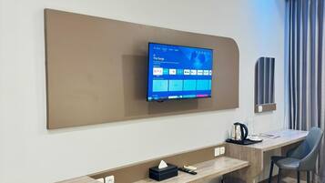 Habitación de hotel moderna con televisión de pantalla plana montada en la pared, un escritorio de madera con un bote de basura, una silla y una pequeña mesa con una cafetera.