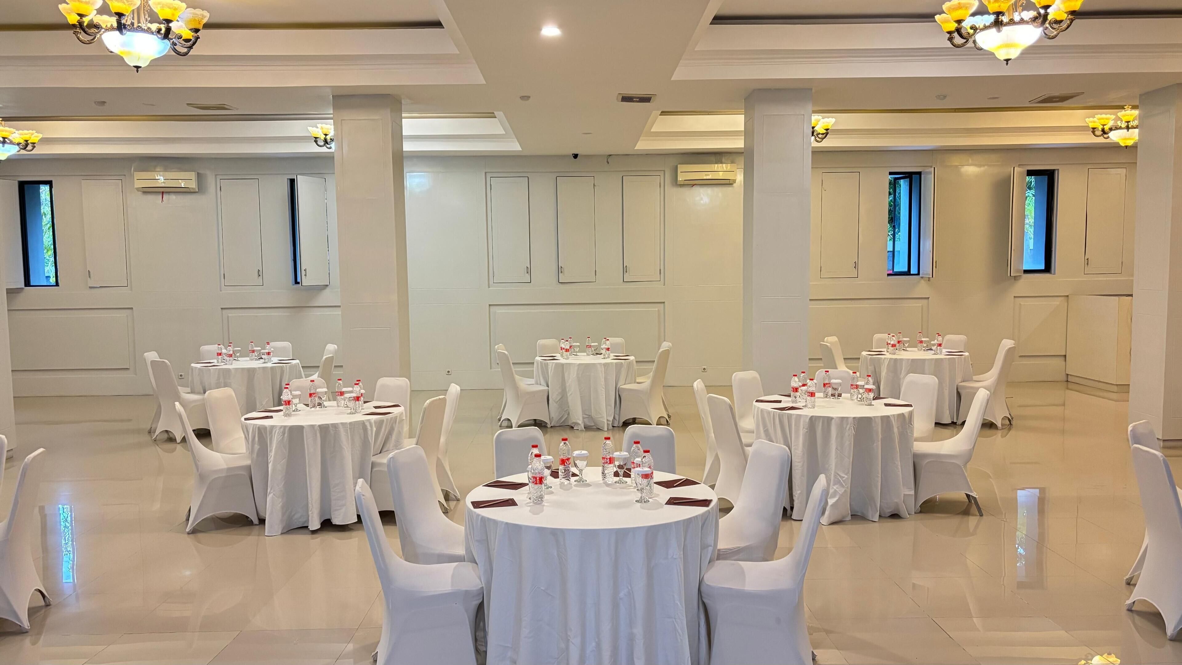 A conference room with multiple round tables, white chairs, and chandeliers.
