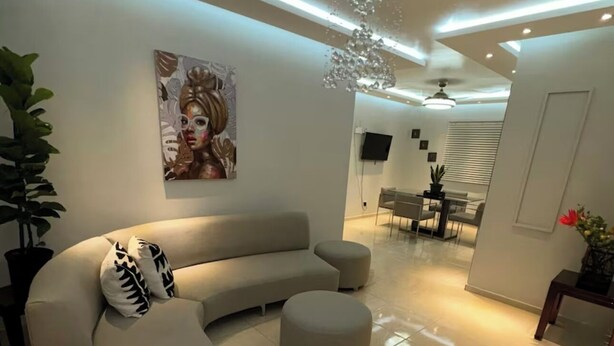 A modern living room with a curved sofa, a round ottoman, a chandelier, and a flat-screen TV mounted on the wall. - 3-bedroom apartment in peaceful San Francisco de Macorís with AC (San Francisco de Macorís)