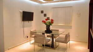 A dining area with a glass table and four chairs, a ceiling fan, a mounted TV, and a window with blinds. - 3-bedroom apartment in peaceful San Francisco de Macorís with AC (San Francisco de Macorís)