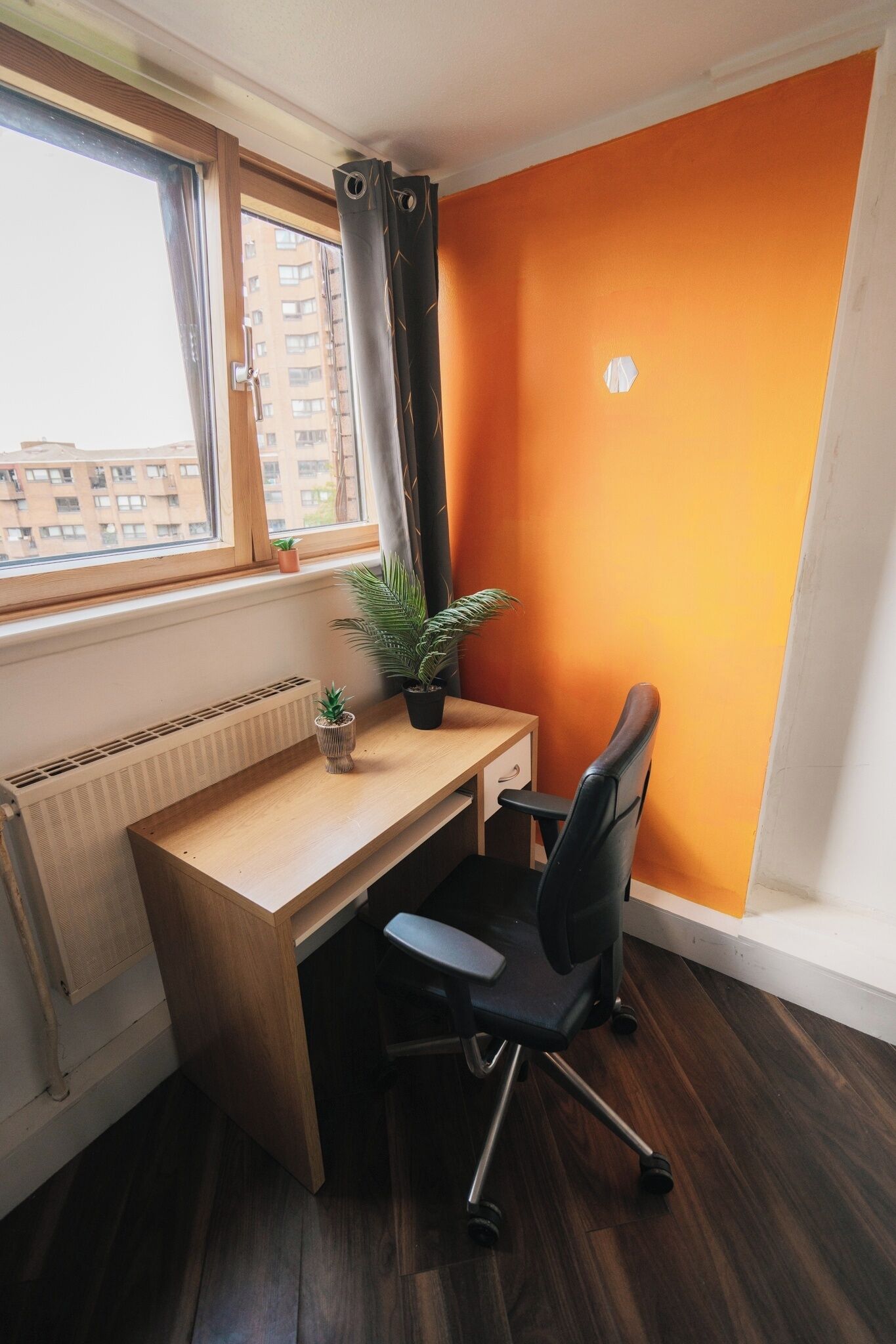 Un pequeño espacio de oficina con un escritorio de madera, una silla de oficina negra y un radiador. Hay dos plantas en macetas en el alféizar de la ventana y se ve una vista de edificios afuera.