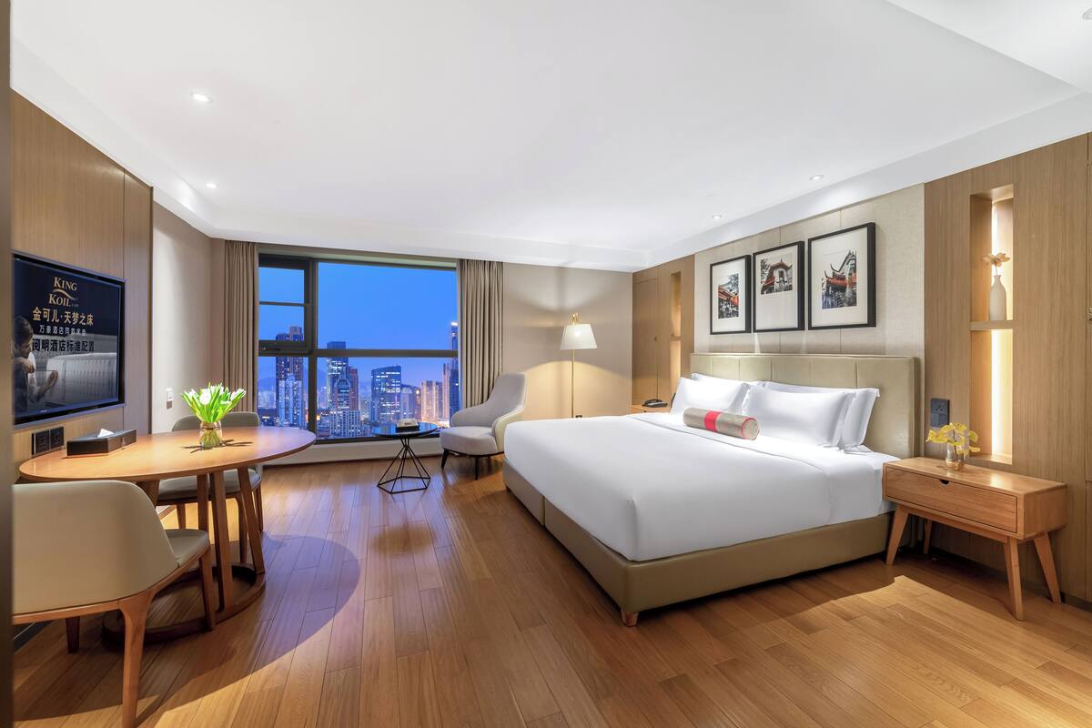 Ein Hotelzimmer mit einem großen Bett, einem Schreibtisch mit Stuhl, einem Fernseher und Blick auf die Stadt.