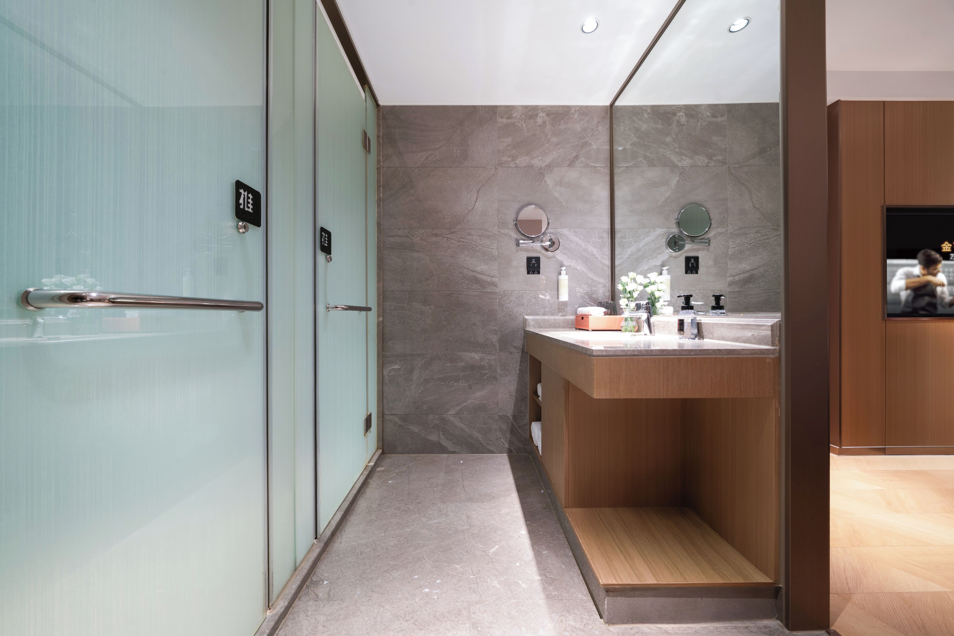 Un baño moderno con puerta de vidrio, tocador de madera y espejo sobre el lavamanos.