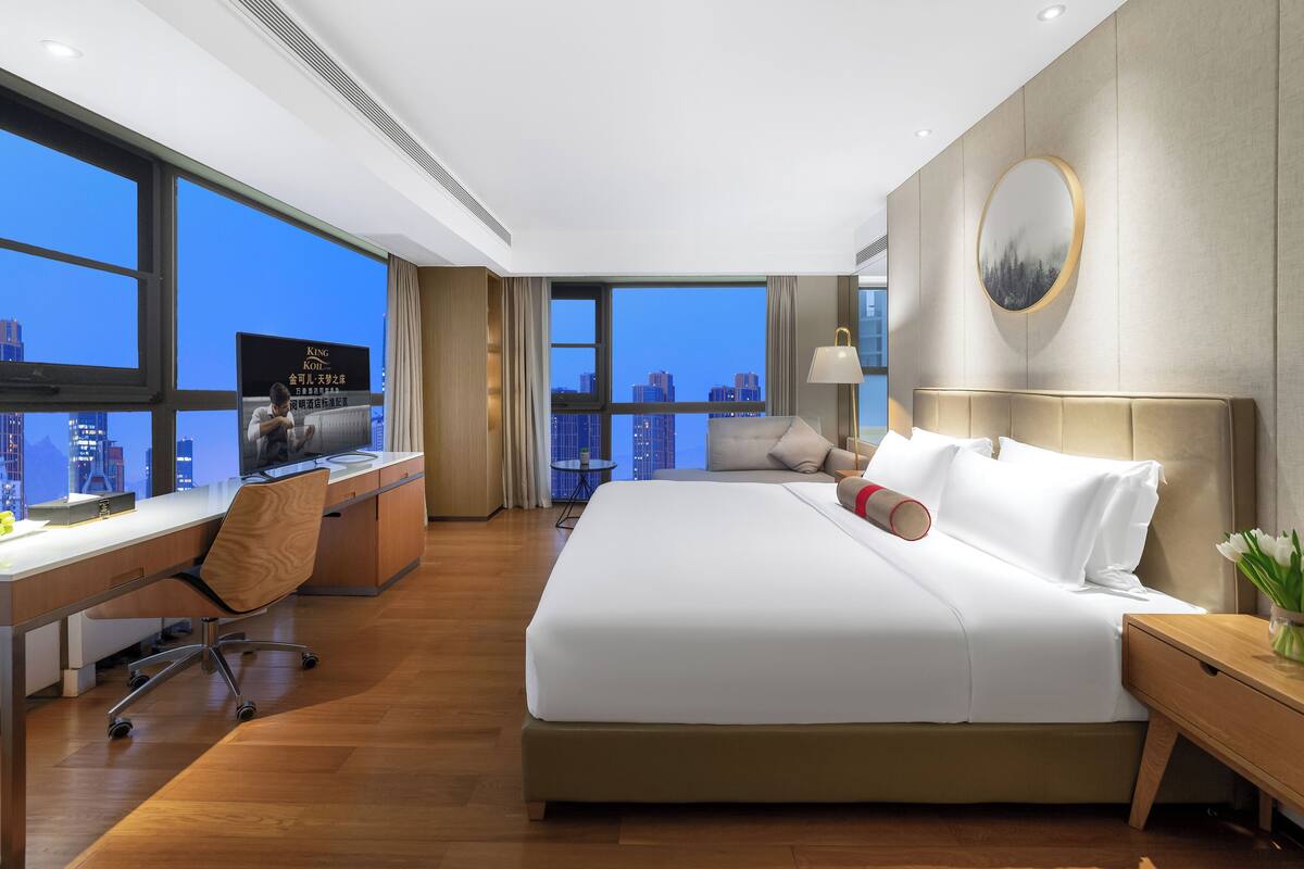 Ein modernes Hotelzimmer mit einem großen Bett, einem Schreibtisch mit Computer und Blick auf die Stadt.