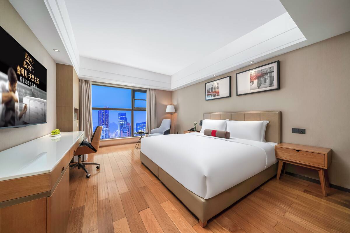 Ein modernes Hotelzimmer mit einem großen Bett, einem Schreibtisch, einem Sessel, einem Nachttisch und einem Fernseher.