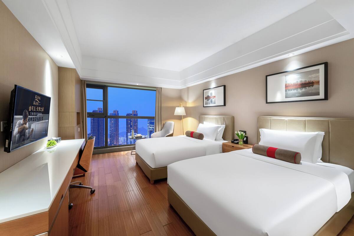 Ein Hotelzimmer mit zwei Betten, einem Schreibtisch, einem Fernseher und einem großen Fenster mit Blick auf die Stadt.