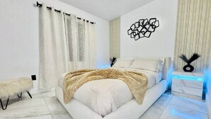 Un dormitorio moderno con una cama grande, cortinas blancas, un adorno en la pared y una mesita de noche con luz azul.