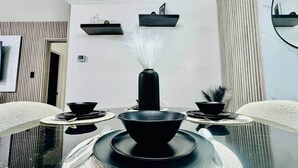 Un comedor con una mesa negra central, cuencos negros y un florero negro con flores blancas. Hay dos estantes negros en la pared sobre la mesa.
