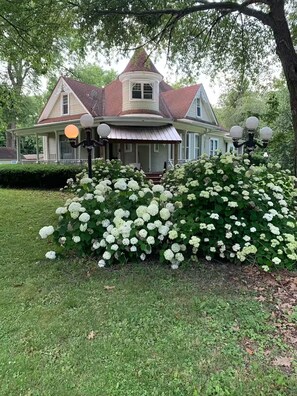A Victorian-style house with a wrap-around porch, a turret, and a large garden with white hydrangeas.