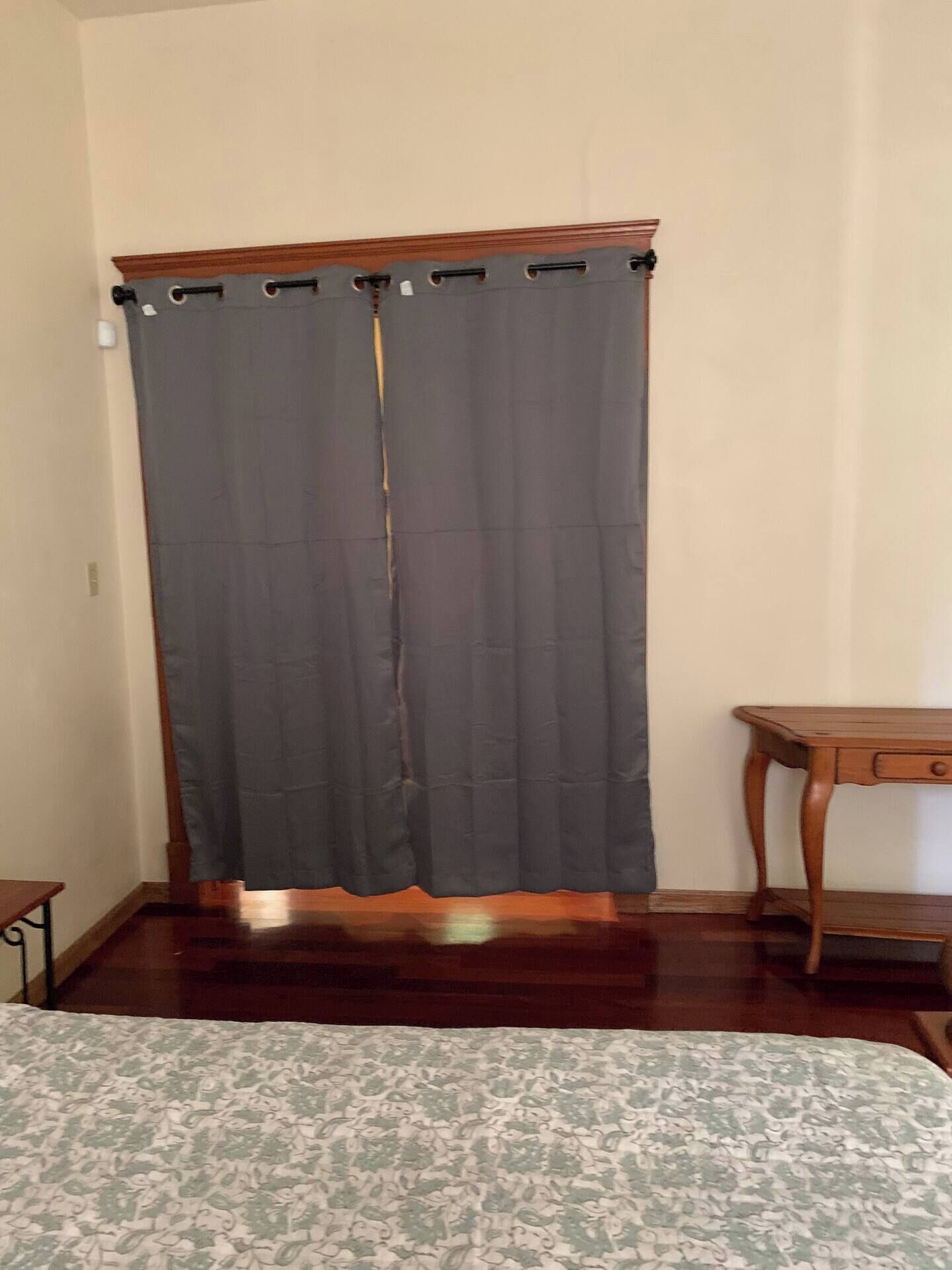 A room with a bed, a wooden nightstand, a small table, and a large window with a curtain.