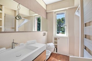 A modern bathroom with a large mirror, white sink, and a window providing a view of trees.