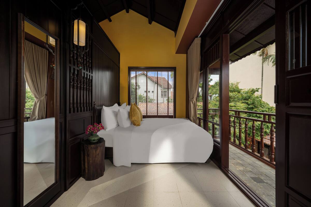 Un dormitorio con una cama grande, cabecera de madera, una mesita con un florero y vista a una casa a través de una ventana.