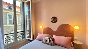 A bedroom with a bed, pillows, a nightstand, a wall-mounted light, and a window with a view of buildings.