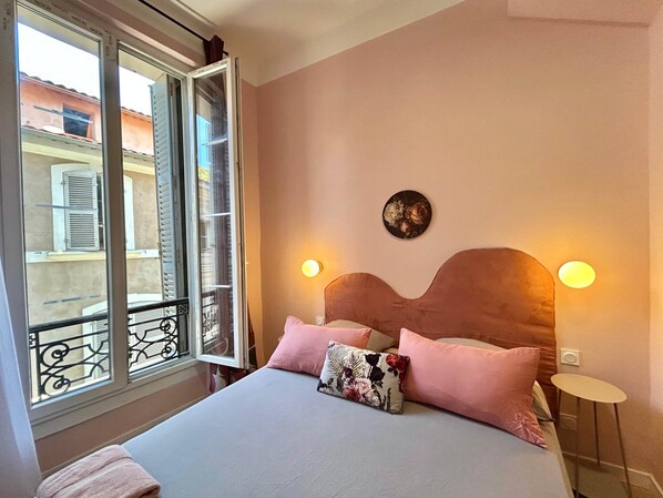 A bedroom with a bed, pillows, a nightstand, a wall-mounted light, and a window with a view of buildings.
