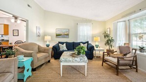 A living room with a beige sofa, a blue sofa, a wooden chair, and a coffee table with a plant. There is a painting on the wall and a window with curtains. - Coastal Cottage Vibes 𓇼 Walk to Beach & Dining (Oak Island)