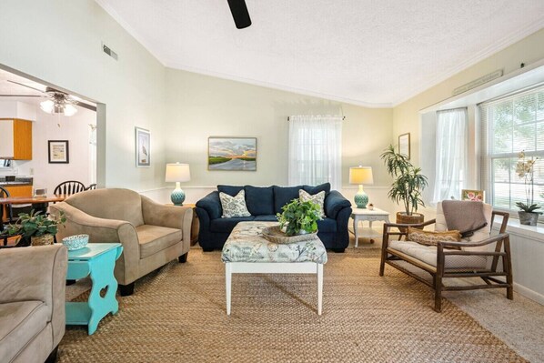 Un salon avec un canapé beige, un canapé bleu, une chaise en bois et une table basse sur laquelle est posée une plante. Il y a un tableau au mur et une fenêtre avec des rideaux.