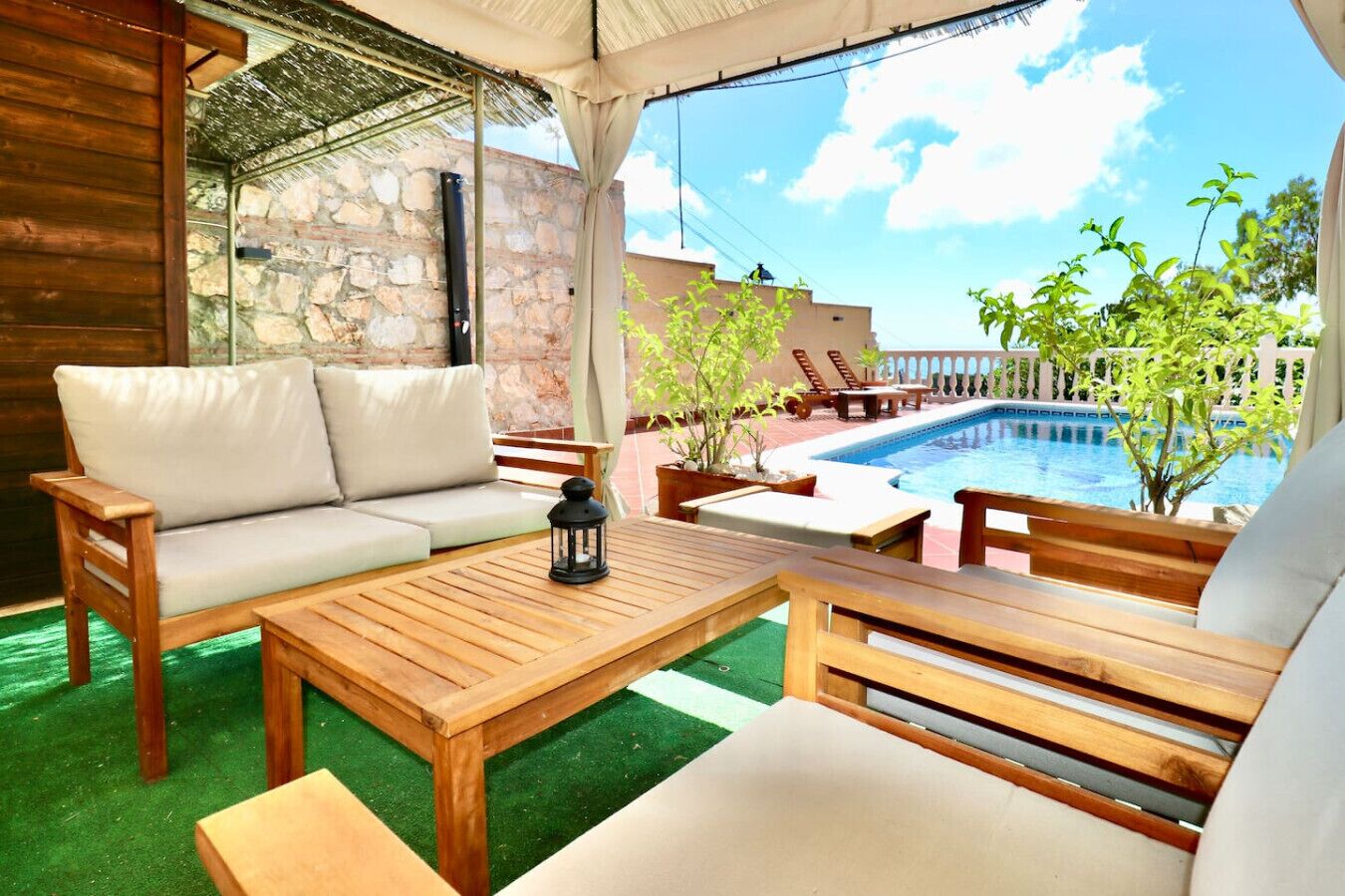 A wooden outdoor seating area with a sofa and tables, a stone wall, and a pool.