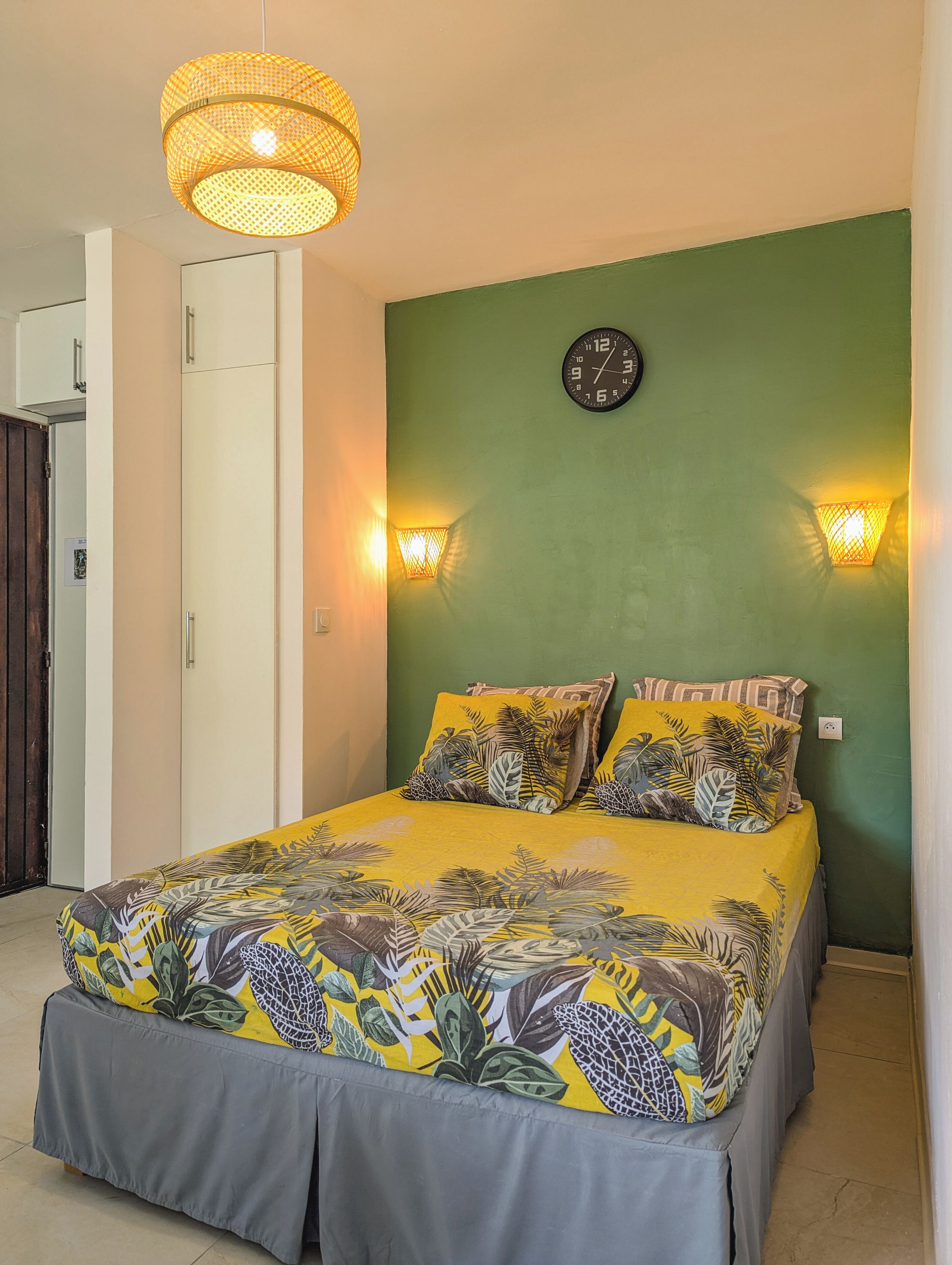 A bed with a tropical leaf-pattern bedspread, a wall clock, and two wall-mounted lights.